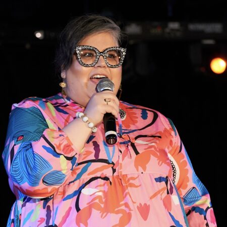 Photo of Issa Kixen giving a speech. She has short dark hair, glasses with large sparkling rims, and a flowing pink and blue shirt.