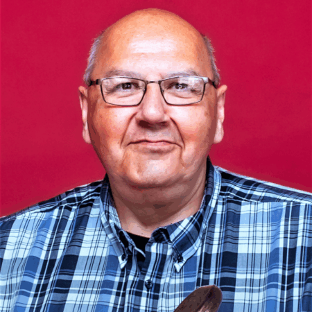 Albert McLeod wearing a blue plaid shirt and glasses smiling in front of a red background.