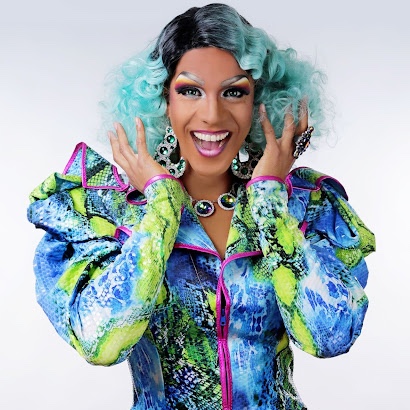 Barbada smiling in a curly teal wig wearing a vibrant blue and green jacket with lavender piping.