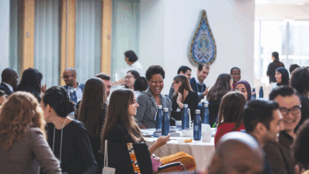 A crowded conference hall with people sitting around tables. In the centre of the picture is a woman smiling to one of her neighbours.