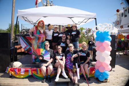 A group f people sitting in a booth at a Pride festival surrounded by pride progress flag. A garland of balloons in blue, pink, and white are arranged like a trans pride flag.