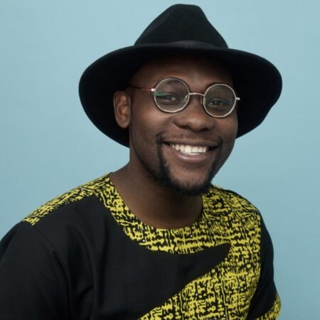 Headshot of Jonah Ssenyange wearing a black fedora, a yellow and black shirt and glasses.