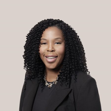 In Leticia's headshot, she's smiling in front of a neutral background with a black blazer and shirt on.